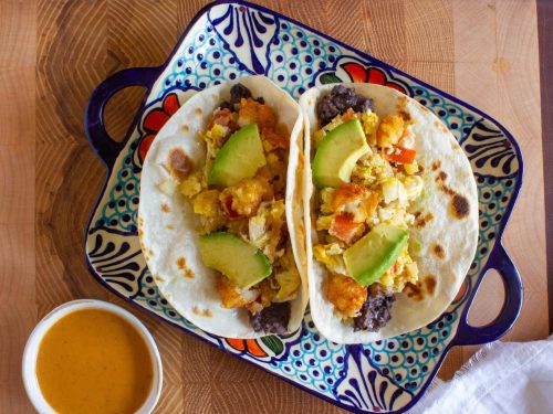 Two tacos filled with avocado, vegetables, and cheese on a decorative plate.